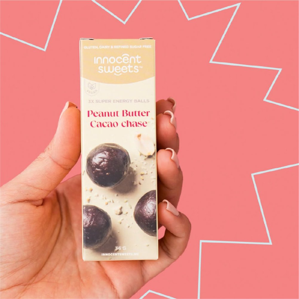 Hand holding a box of 'Peanut Butter Cacao Chase' by 'innocent sweets' against a pink background